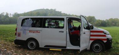 Der Bürgerbus Niedenstein steht vor einem Wald auf einem Feld. Die Beifahrer Tür ist offen und mehrere menschen sitzen in dem Bus drinnen.