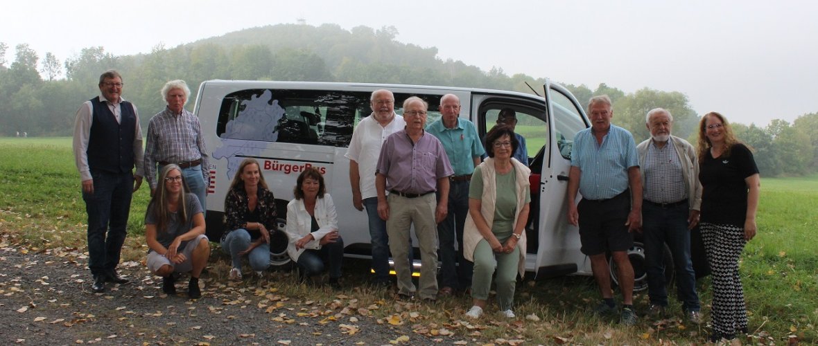 DerBürgerbus Niedenstein steht auf einem Feld vor einem Wald davor und darin stehen oder sitzen ein paar Menschen.