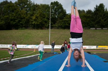 Auf dem Bild ist ein Mädchen mit einer rosanen Hose und weißem Top und roten Haaren die einen Handstand macht auf einer blau weißem Matte. Im hintergrund sind mehrere kinder mit buten Anziehsachen die rennen. Der boden hat rote Steine.
