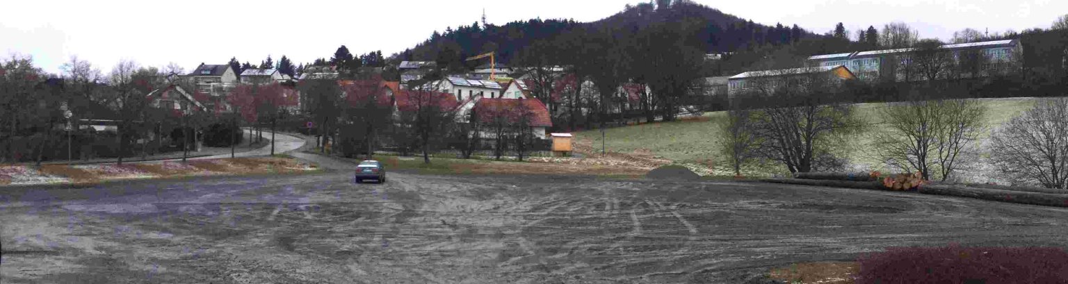 geschotteter Platz mit einem parkenden Auto im Hintergrund Wohnbebauung, Hessenturm und Grundschule